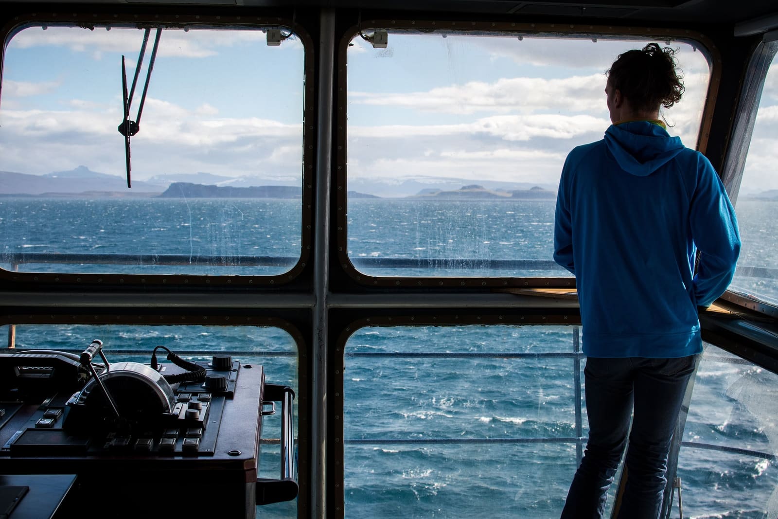 A person on the bridge looking out through the windows at a choppy sea and a distant coastline.