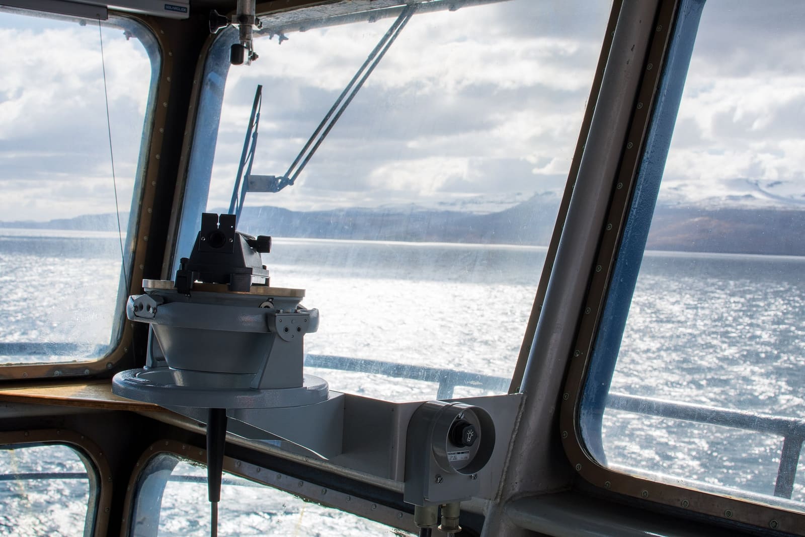 The view from a ship’s bridge window: bright sea, mountains on the horizon and a searchlight on the bulwark.
