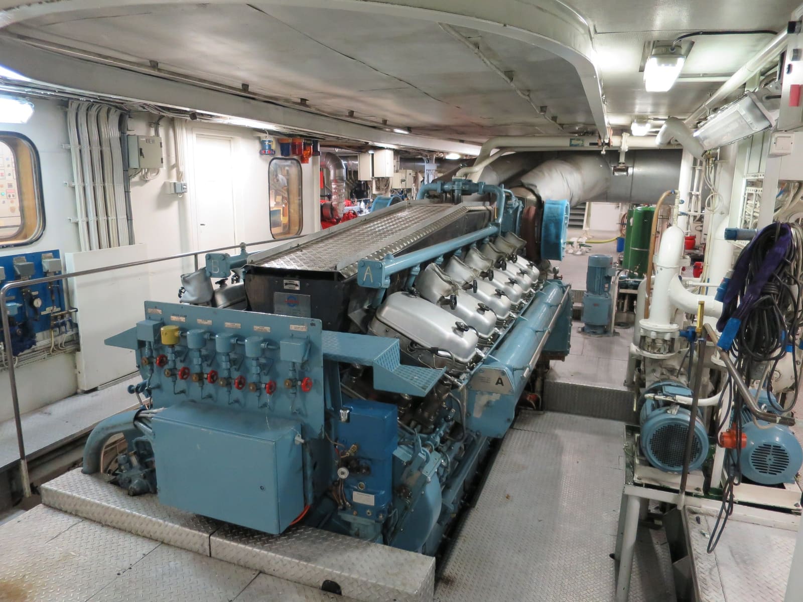 A clean engine room with a large blue diesel engine, pipework and pumps under fluorescent light.