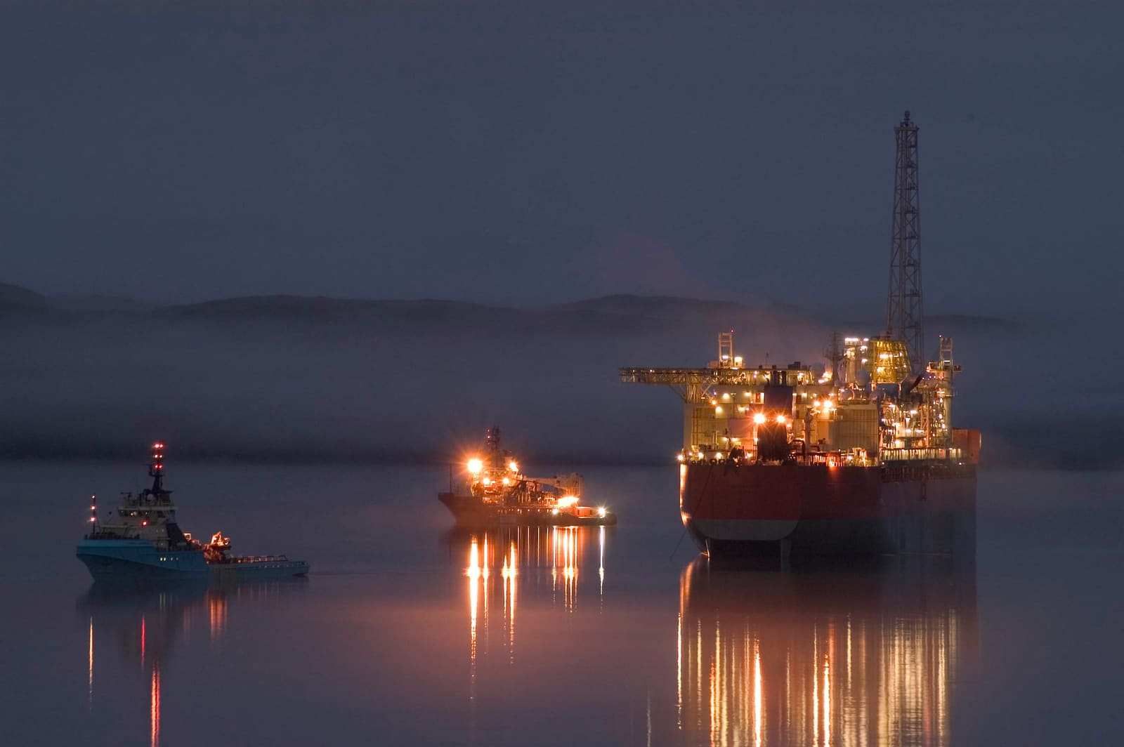 A production ship and two offshore support vessels lit up on still water at dusk, their lights reflected in the sea.