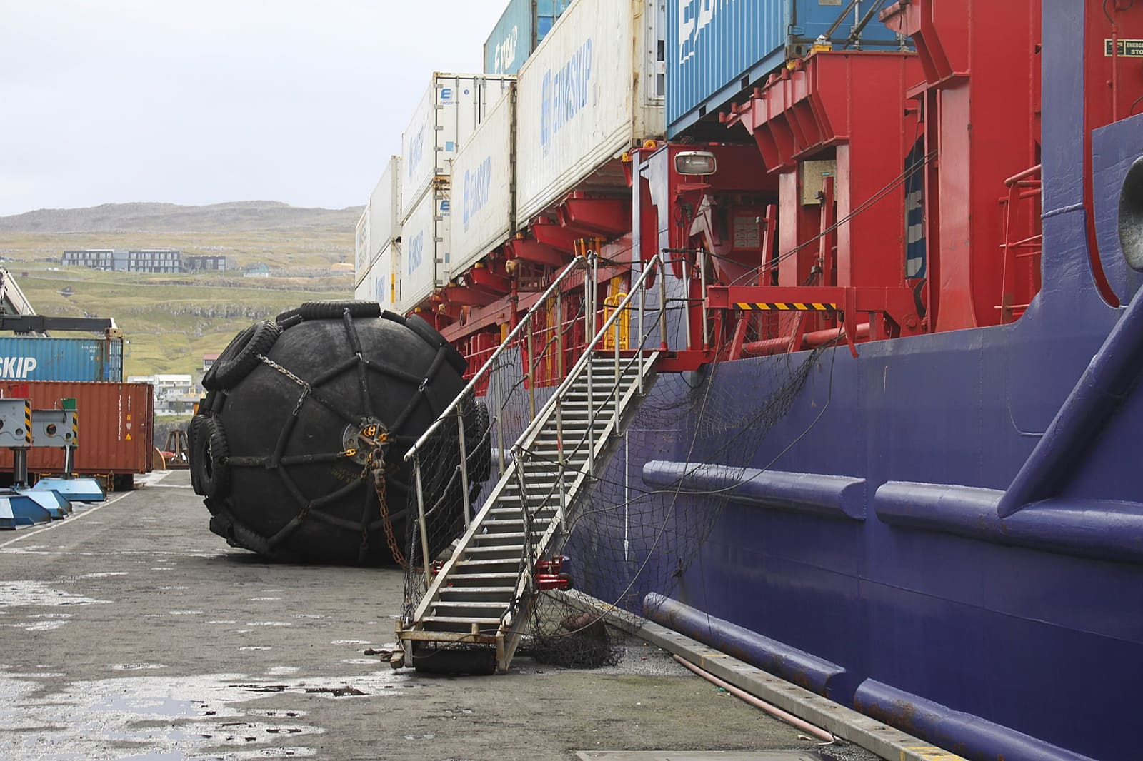 A ship’s gangway rigged down to the quay beside a large fender, with the hull rising above.