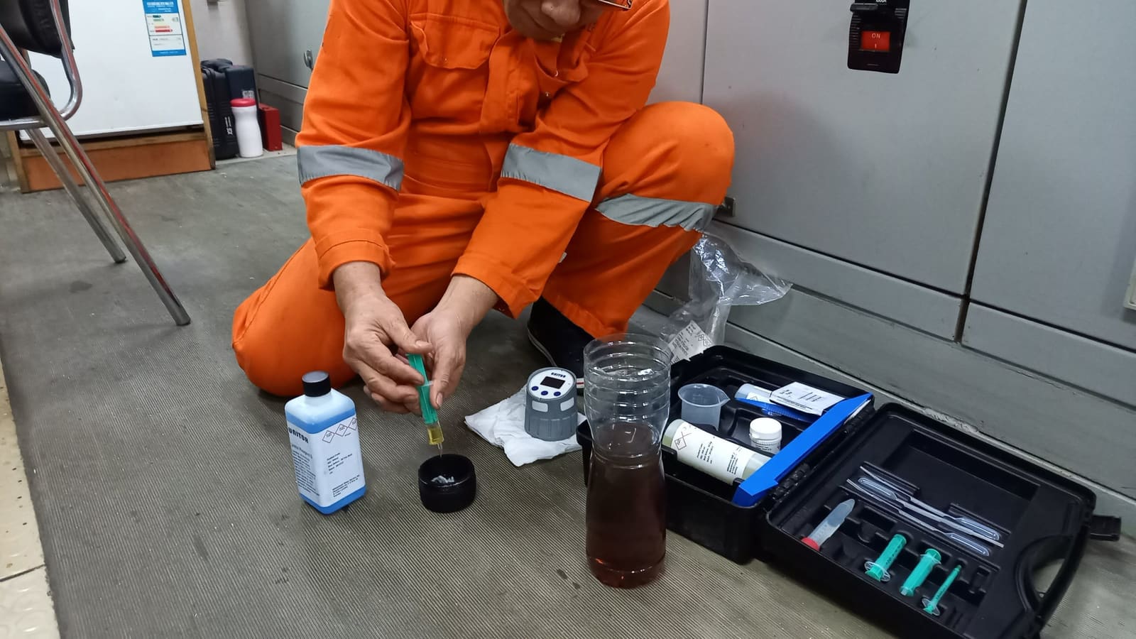 A crew member in an orange coverall kneeling on deck to test an oil sample with a test kit.