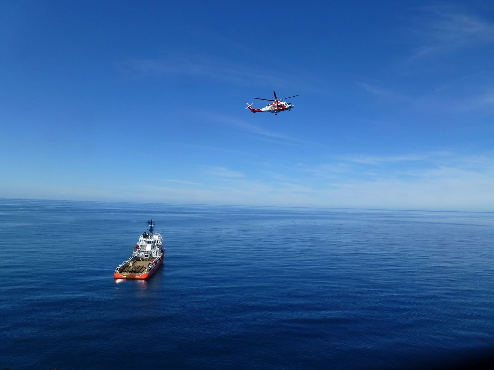 An offshore support vessel on deep blue open sea, seen from a rig, with a helicopter passing overhead.