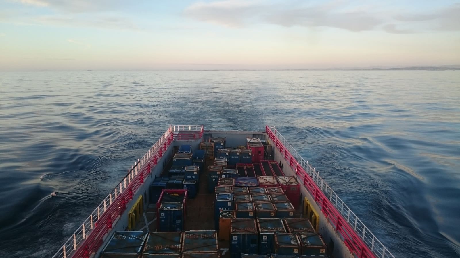 The cargo deck of a platform supply vessel, loaded with containers, heading out over a calm sea at dusk.