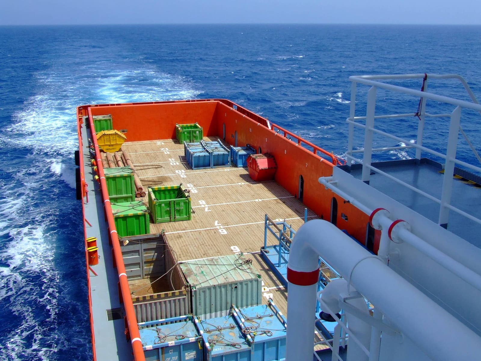 Looking aft over a supply vessel’s cargo deck with containers and baskets, the wake white on a deep blue sea.