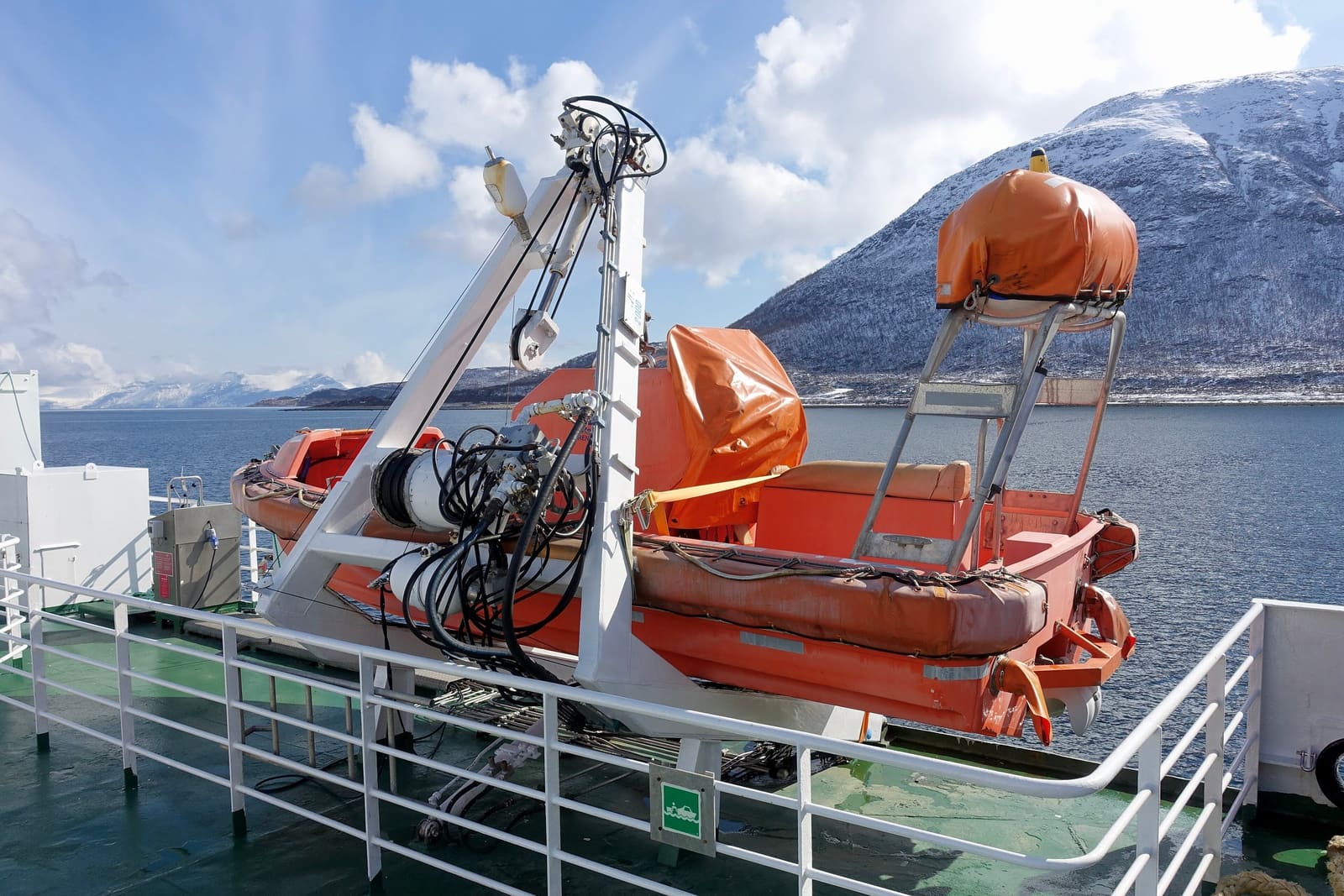 An orange rescue boat in its davit on a ferry’s deck, with snow-capped mountains across a Norwegian sound.