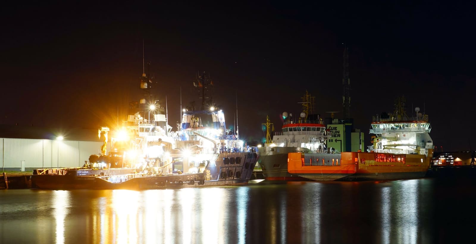 Offshore vessels alongside at night under working lights, reflected in the harbour water.